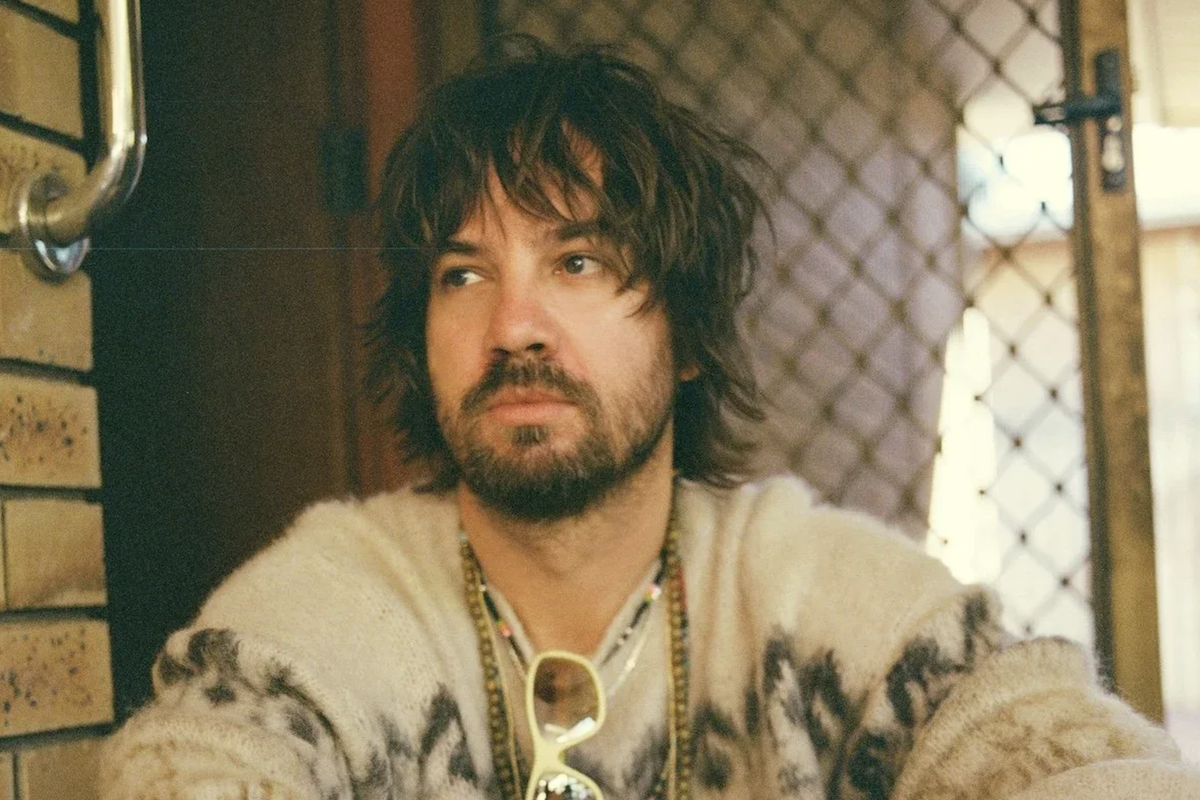 Kevin Parker of Tame Impala wearing a patterned sweater, beaded necklaces, and sunglasses indoors in a casual portrait.